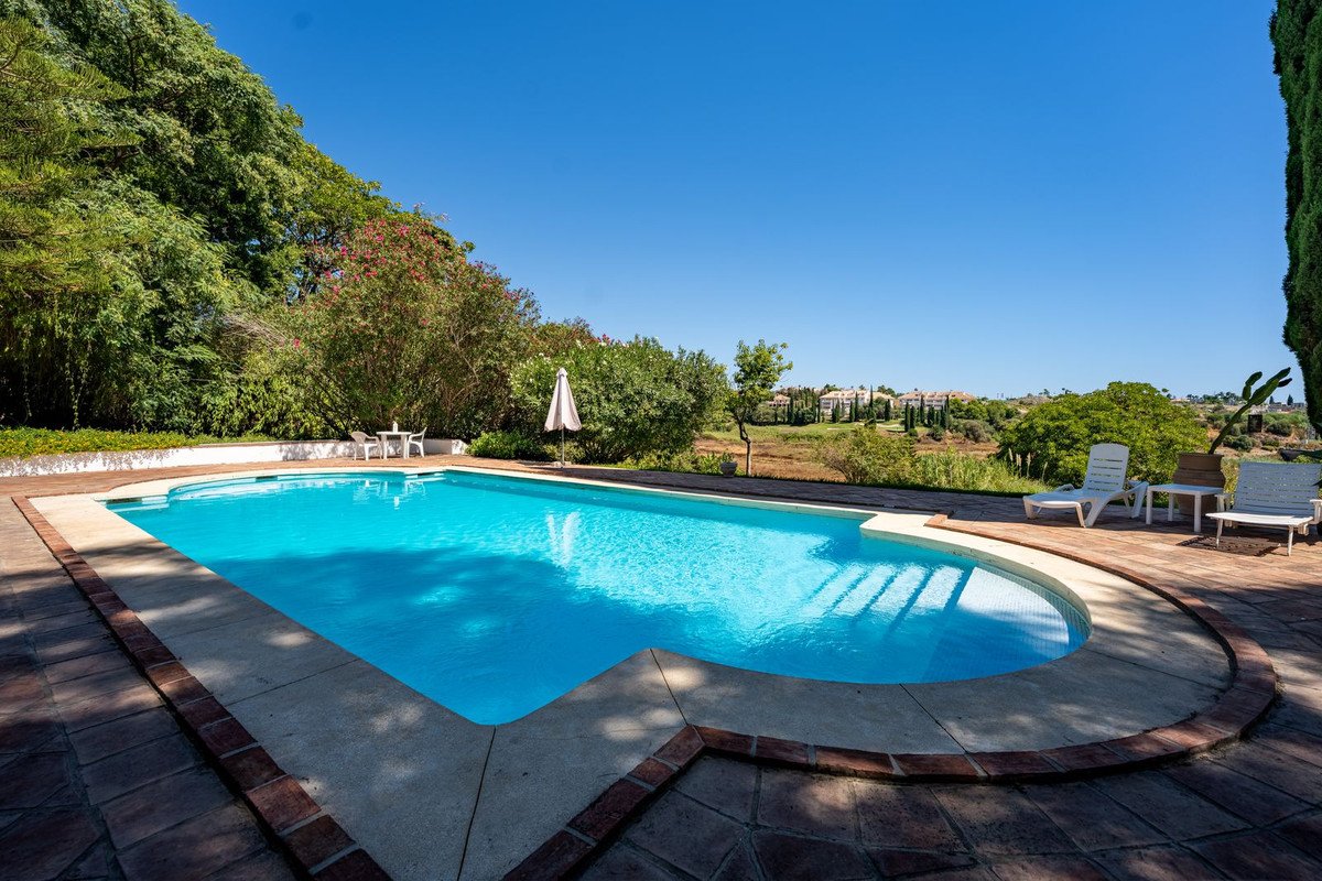 A large, clear blue swimming pool with sun loungers and distant villas under a bright blue sky.