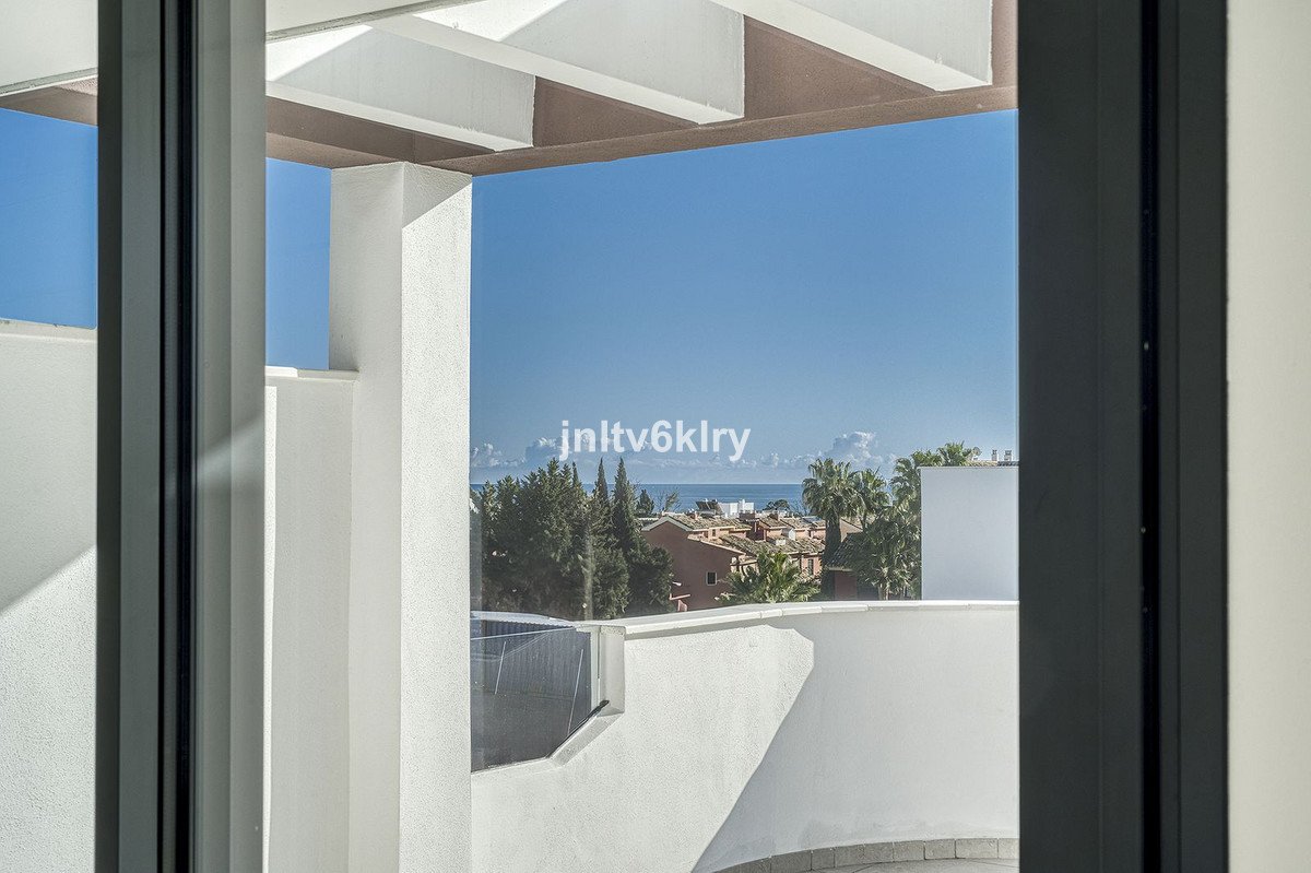 View from a balcony with white railings, overlooking rooftops, palm trees, and the sea under a clear blue sky.
