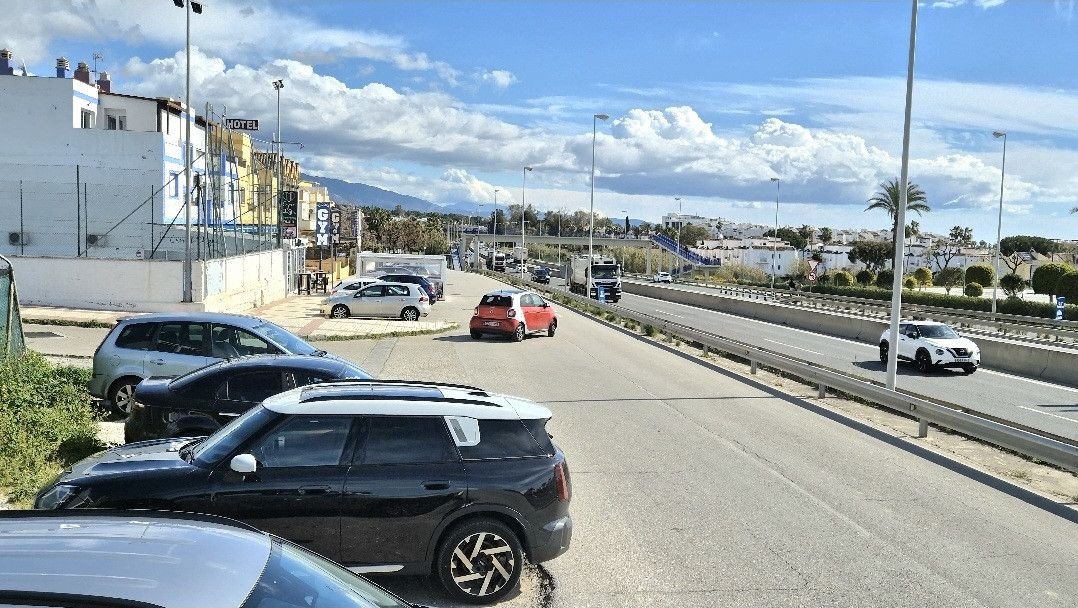 Street view showing multiple cars parked on the side of a road with buildings and a highway in the background under a cloudy sky.