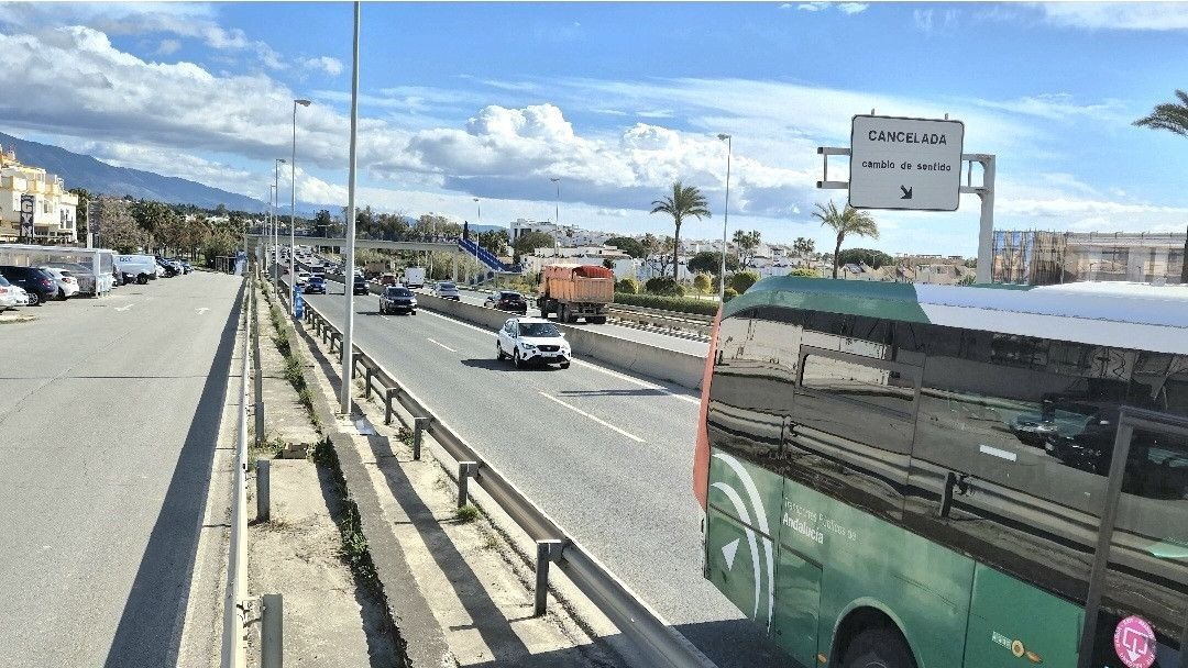 View of a highway with traffic, a bus in the foreground, and buildings and mountains in the background under a cloudy sky.