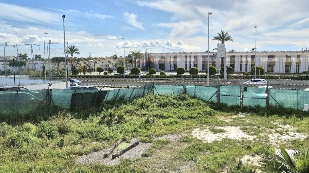 View of a tennis court with a building and palm trees in the background under a cloudy sky.