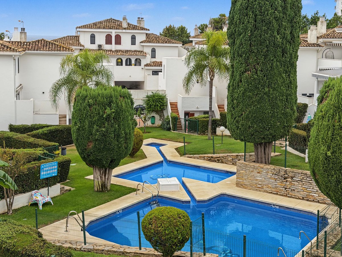 A view of a shared swimming pool complex with lush green gardens and modern white buildings in the background.