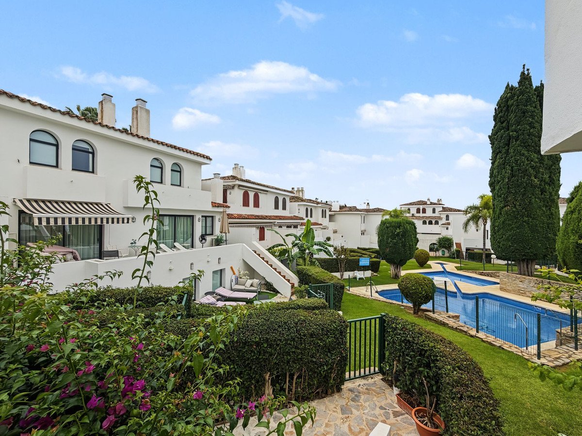 White stucco townhouses with a swimming pool and green garden in a sunny courtyard with blue sky and clouds.