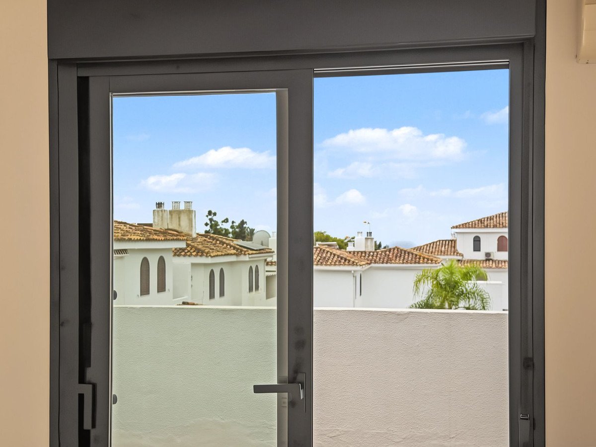View of Mediterranean rooftops and palm trees through a glass door on a sunny day with blue sky and clouds.