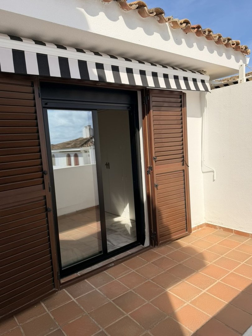 A sunny terrace with terracotta tiles, a sliding glass door, brown shutters, and a black and white striped awning overhead.