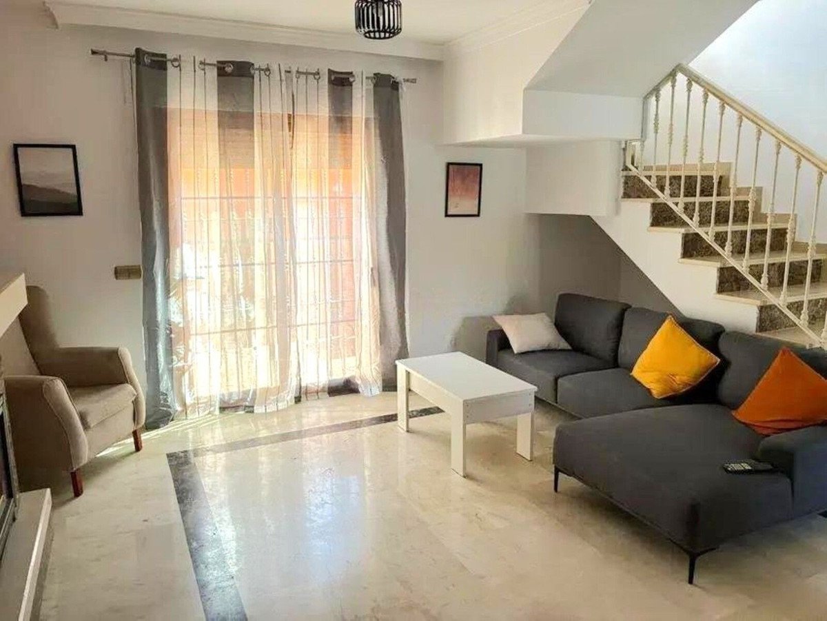 A spacious living room with a sectional sofa, coffee table, armchair, and stairs leading to the upper floor. Large windows with sheer curtains let in natural light.