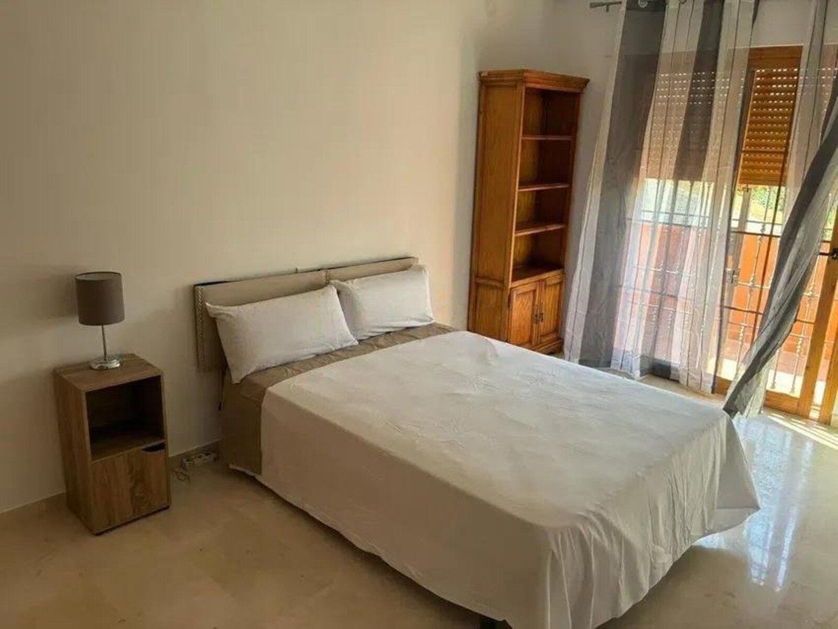 Bedroom with a bed, bedside table, wooden bookcase, and large windows with natural light streaming in.