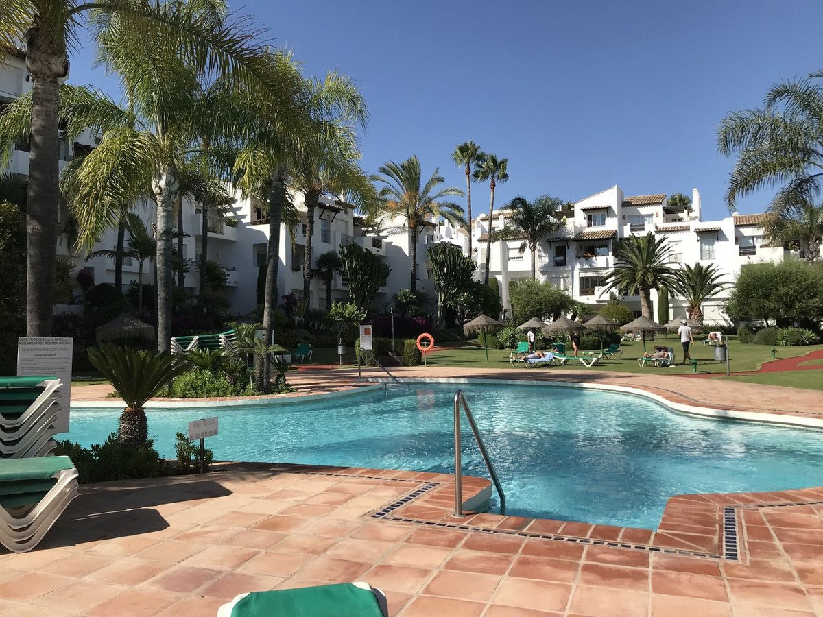 Resort pool area with palm trees, sun loungers, and white apartment buildings under a clear blue sky.