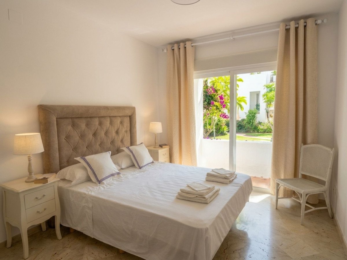 A bedroom with a large bed, tufted headboard, and a sliding glass door leading to a garden with flowers.