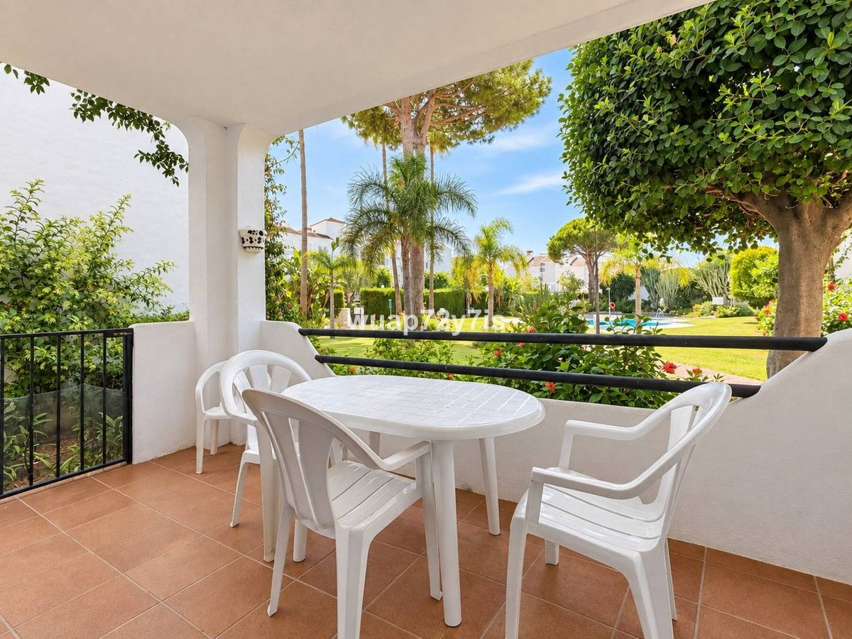 Terrace with white table and chairs overlooking a garden and swimming pool on a sunny day.