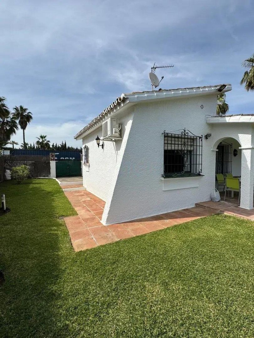 White house exterior with a green lawn, tiled pathway, and a covered terrace with chairs.
