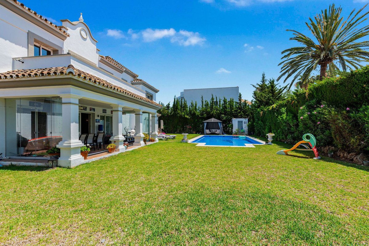 Spacious villa backyard with a swimming pool, green lawn, covered patio, and a palm tree under a clear blue sky.