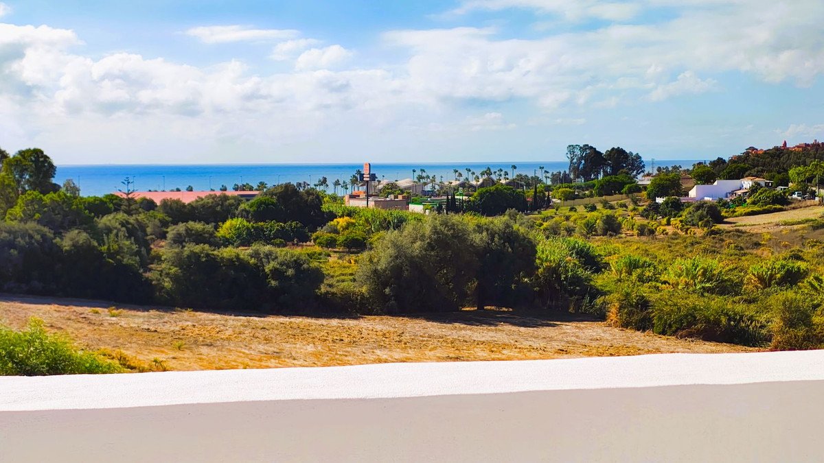 A panoramic view of the sea beyond a green countryside with trees and distant white villas under a blue sky.