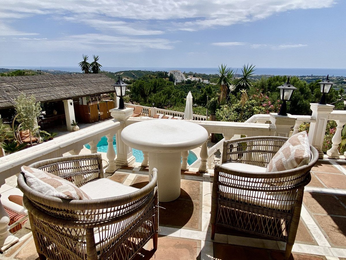 Rattan chairs on a terrace overlooking a pool, lush greenery, and the ocean under a partly cloudy sky.