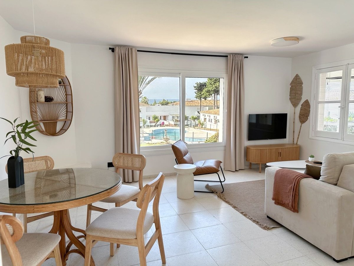 Bright living and dining area with rattan furniture, offering a view of the pool and distant sea.