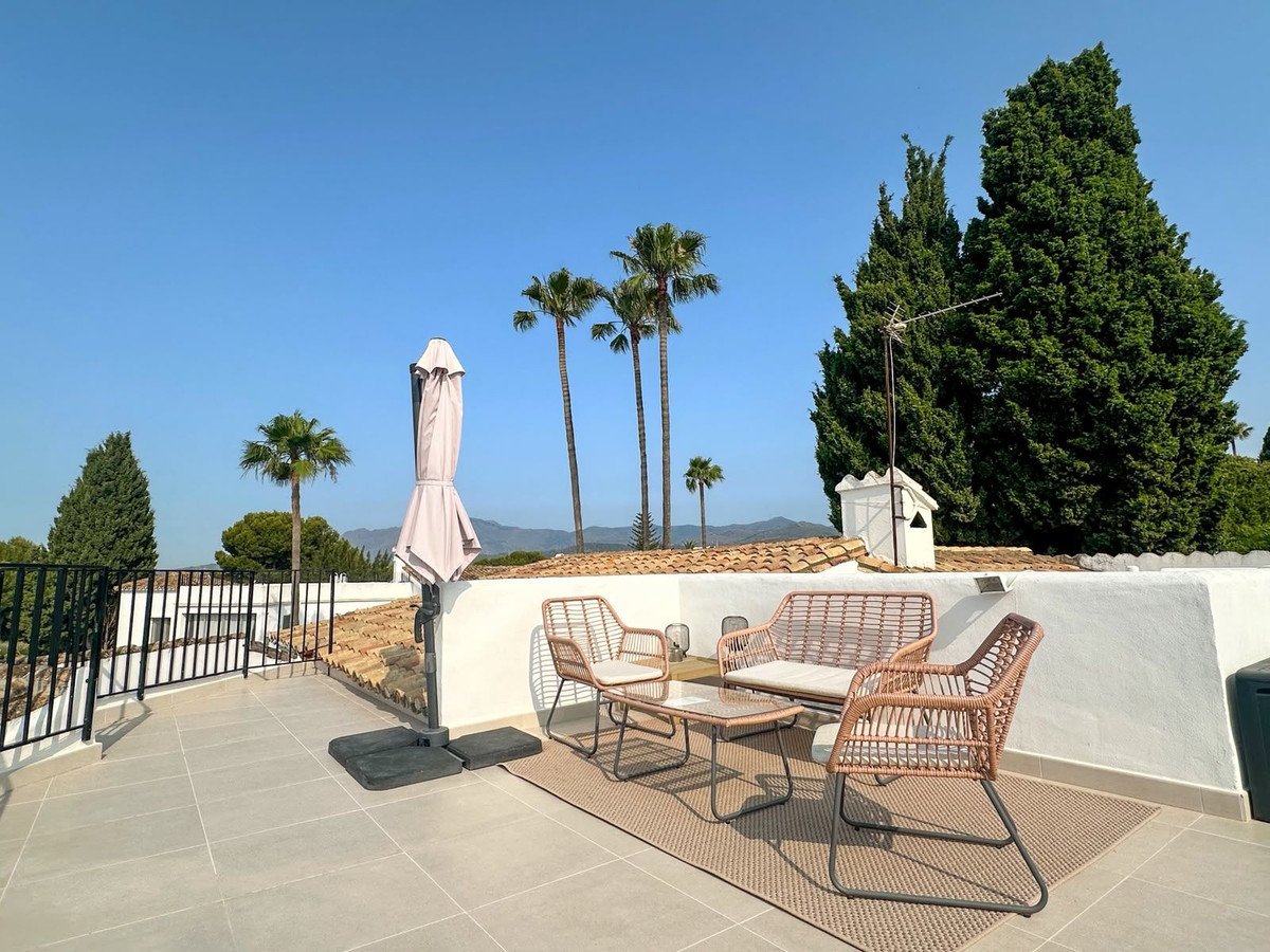 A sunny terrace with rattan seating, umbrella, and a view of palm trees and distant mountains.