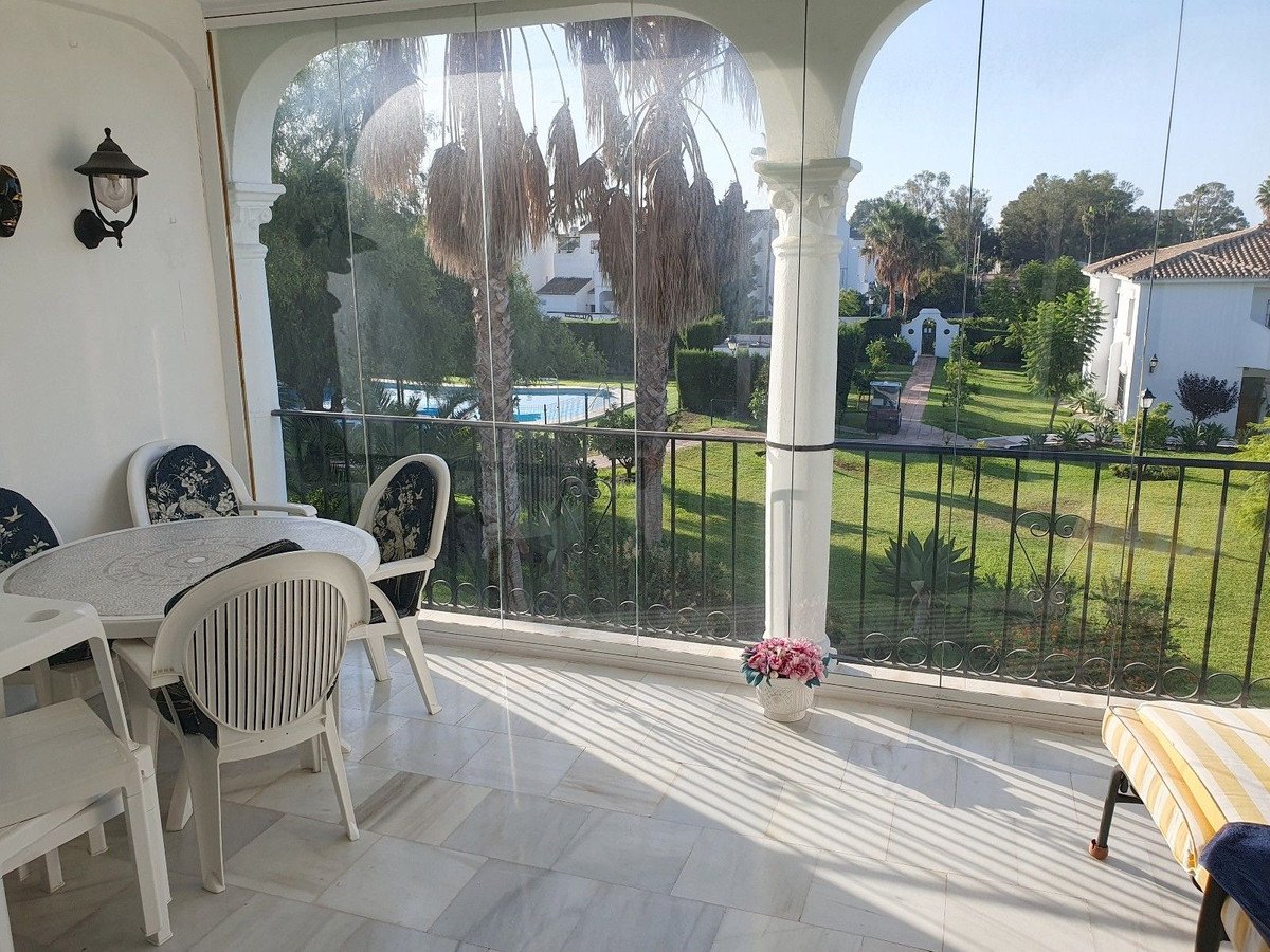 Enclosed patio with table and chairs overlooking a lush garden and swimming pool, with palm trees and white buildings.
