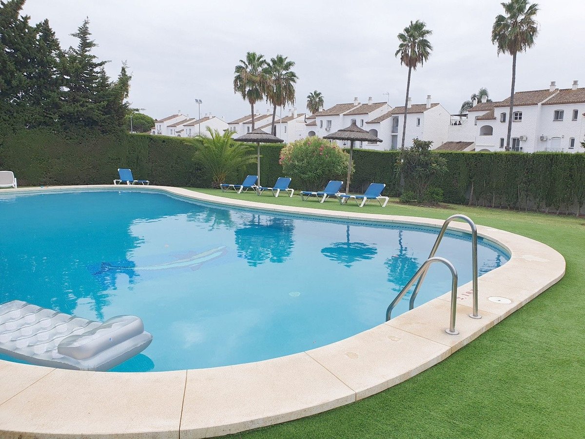 A clear blue swimming pool with loungers and umbrellas, surrounded by green grass and apartment buildings.