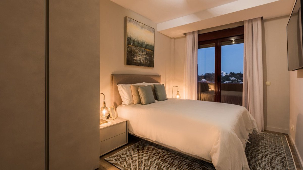 A cozy bedroom with a double bed, bedside tables with lamps, and a large window with a view of the city lights at dusk.