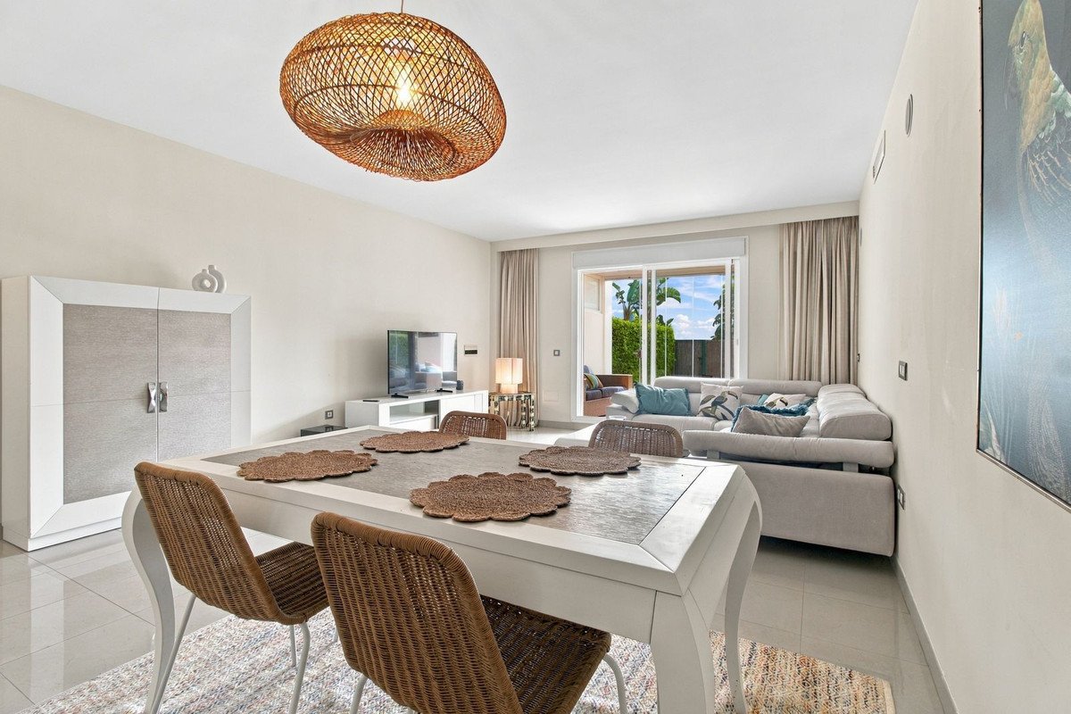 Modern dining and living area with a large sofa, white dining table, wicker chairs, and a view of a lush patio through glass sliding doors.