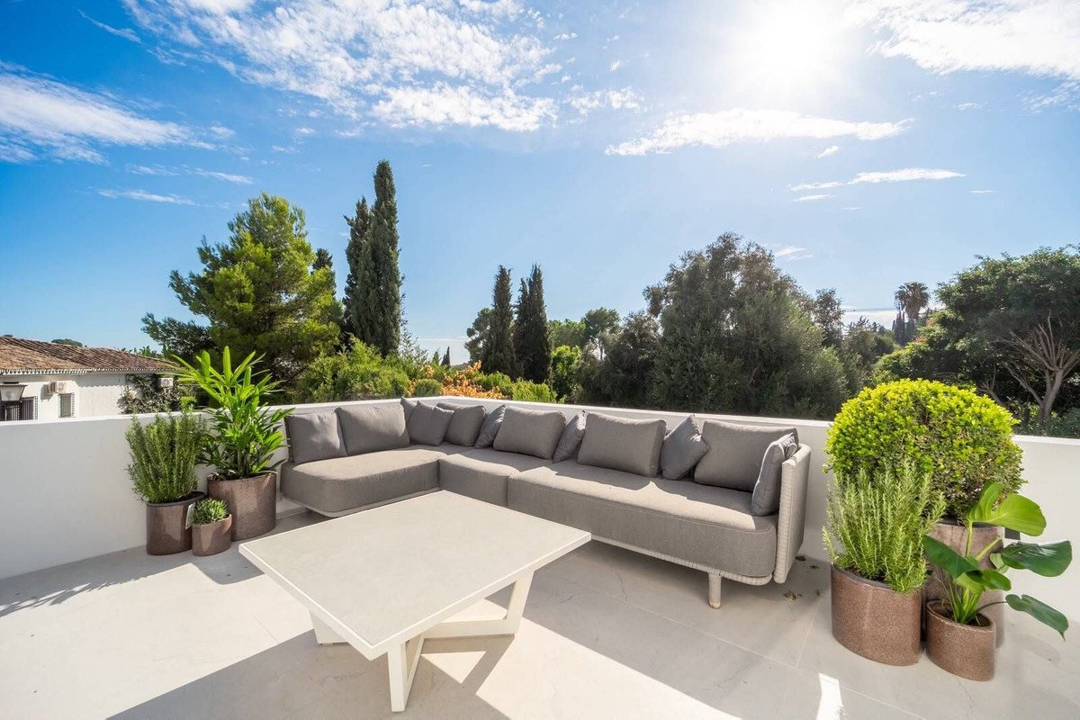 Modern outdoor lounge area with a sectional sofa, coffee table, and potted plants, offering a scenic view of lush greenery under a bright sky.