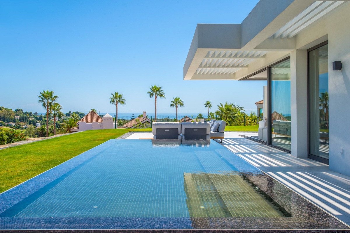 Modern villa with infinity pool overlooking the sea, surrounded by palm trees and manicured lawn. Outdoor seating area visible.