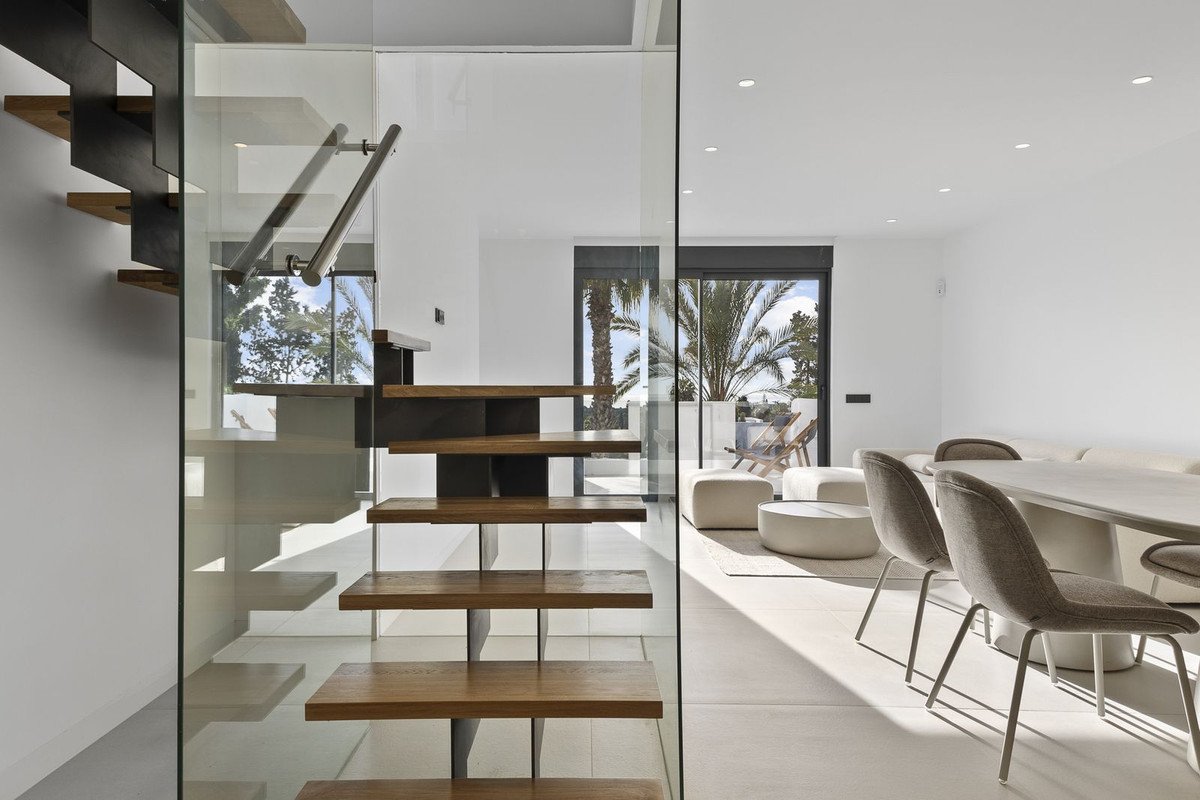 Modern floating staircase with glass railing adjacent to a bright living area with outdoor patio view and palm trees.