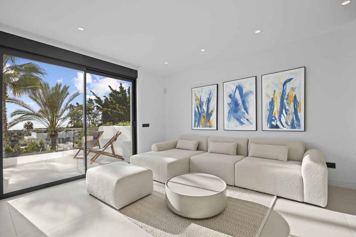 Modern living room with a large sectional sofa, ottoman, round coffee table, and abstract art, opening to a balcony with palm trees and ocean views.