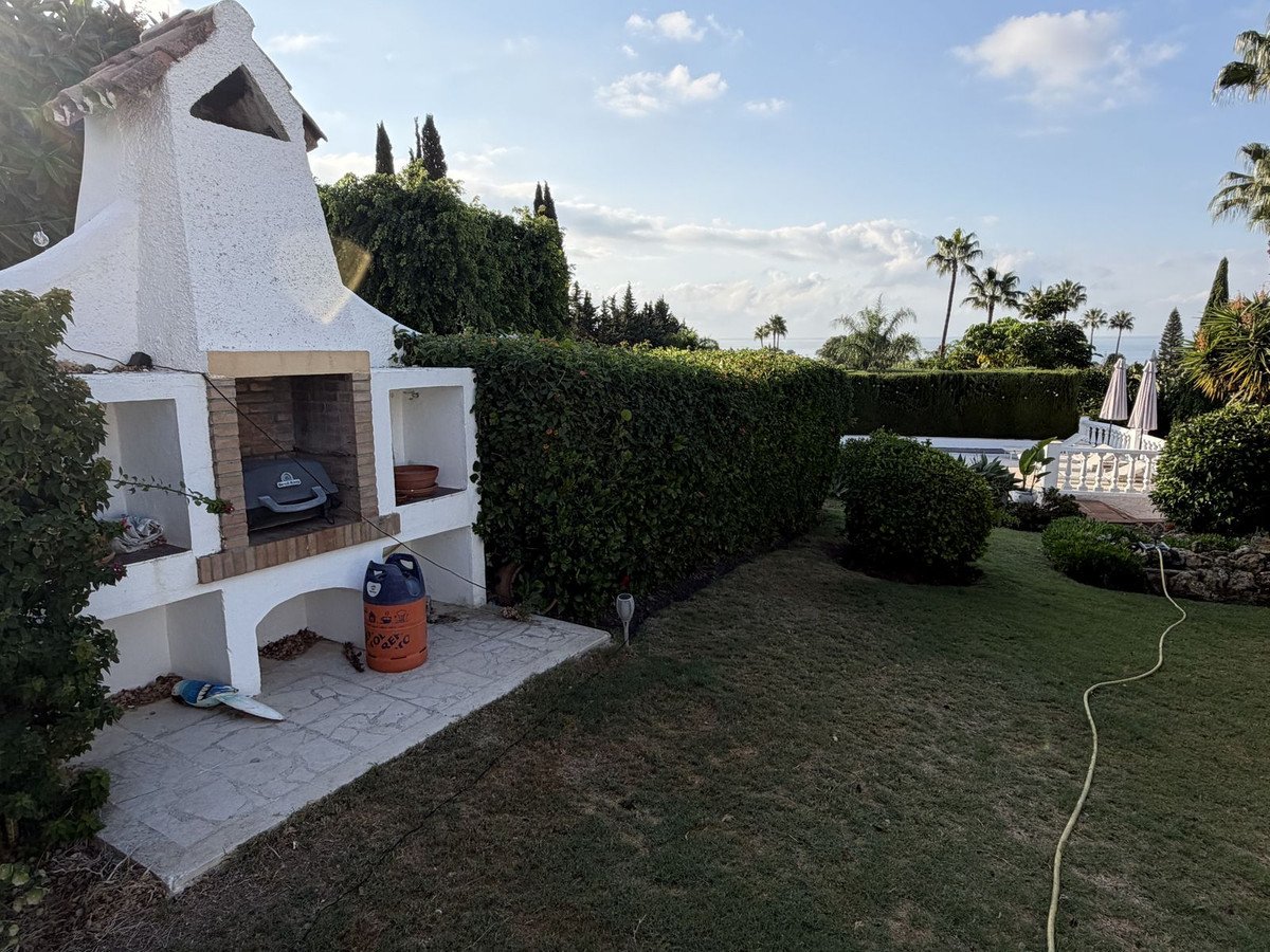 Outdoor BBQ area with a built-in grill, leading to a garden with a swimming pool and distant sea view.
