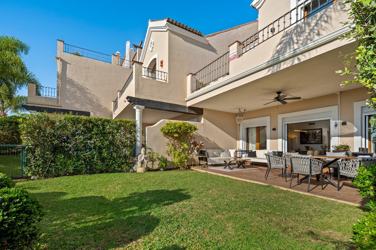 Terrace with outdoor seating and dining areas, overlooking a green lawn and a beautiful house exterior under a clear blue sky.
