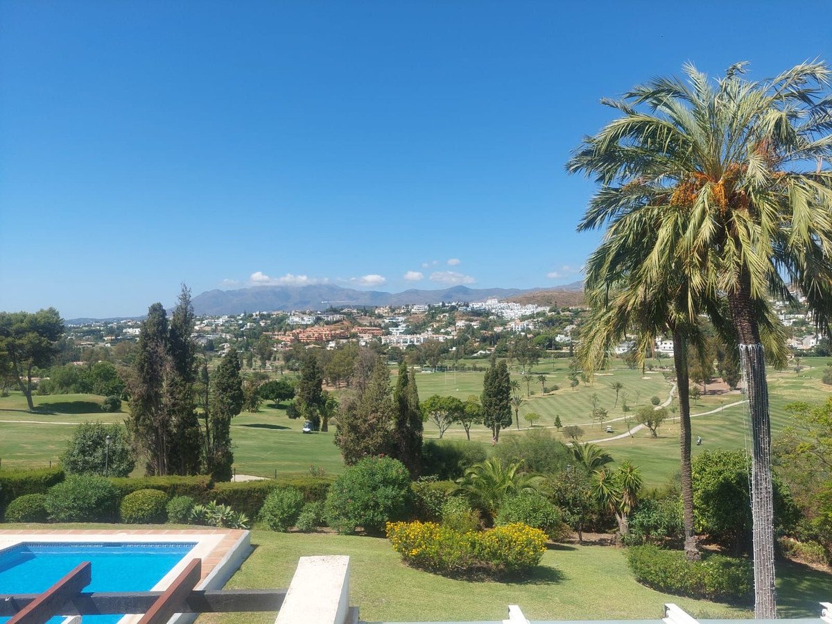 Balcony view of a swimming pool, lush gardens, a golf course, and distant hills with palm trees.