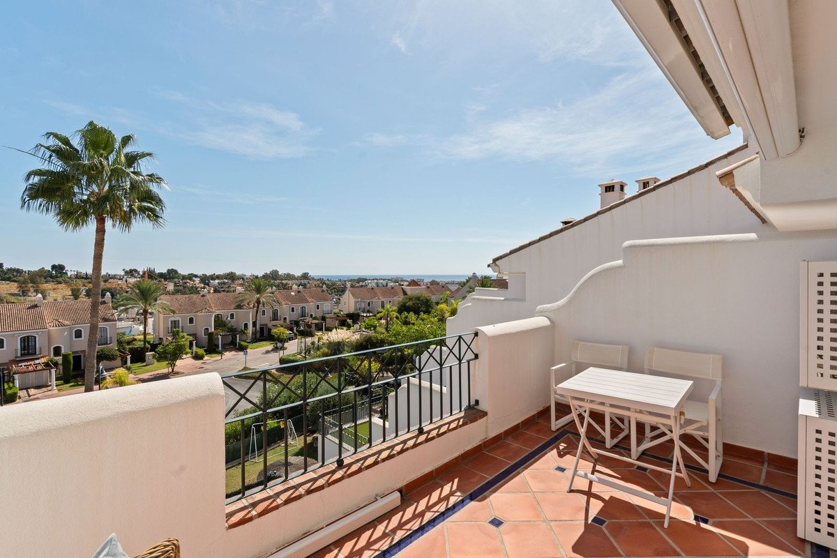 A balcony with a white table and chairs overlooking a residential area with palm trees and distant sea view under a clear blue sky.