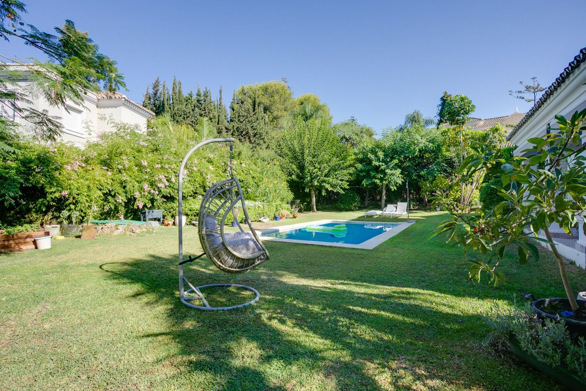 A lush green garden with a swimming pool, lounge chairs, and a hanging swing chair under a clear blue sky.