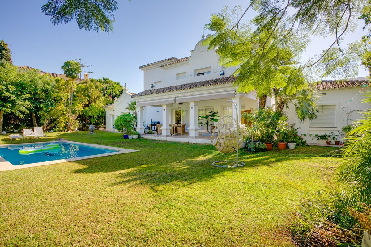 A white villa with a swimming pool, lush green garden, covered patio, and hanging chair on a sunny day.