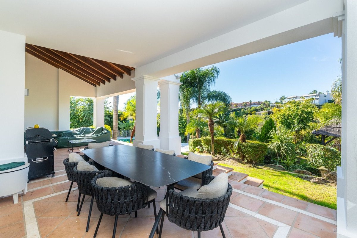 Covered patio dining set with grill and seating, overlooking a lush garden with a pool and distant houses on a hill.