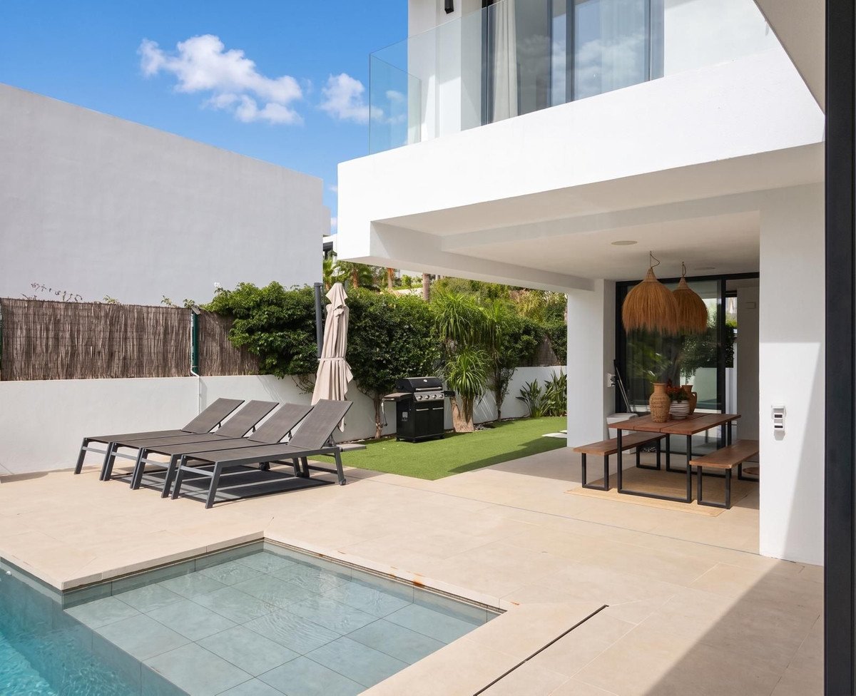 Modern outdoor area with a swimming pool, sun loungers, dining table, barbecue grill, and lush green garden.