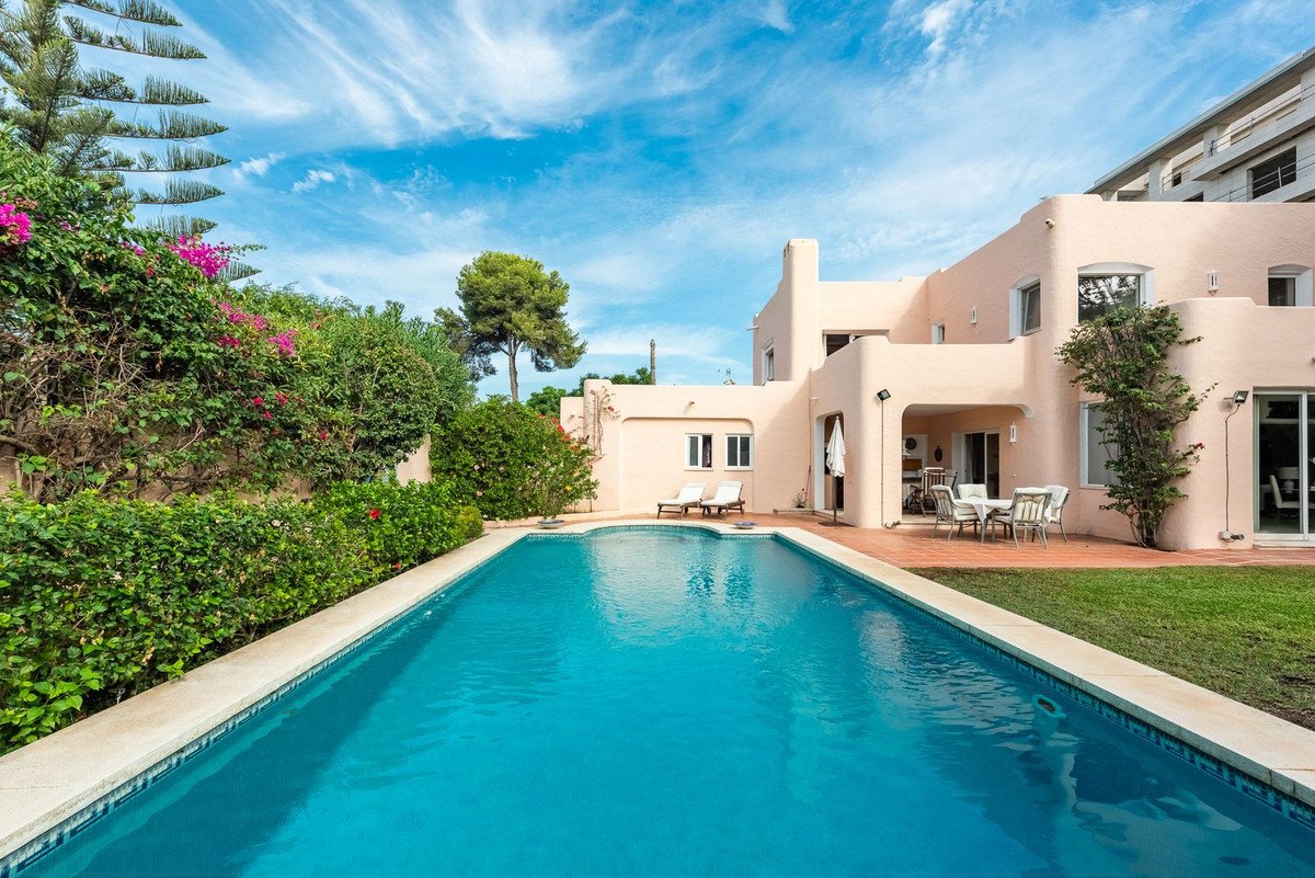 A large rectangular swimming pool in front of a light peach villa with an outdoor dining area and lush green landscaping.