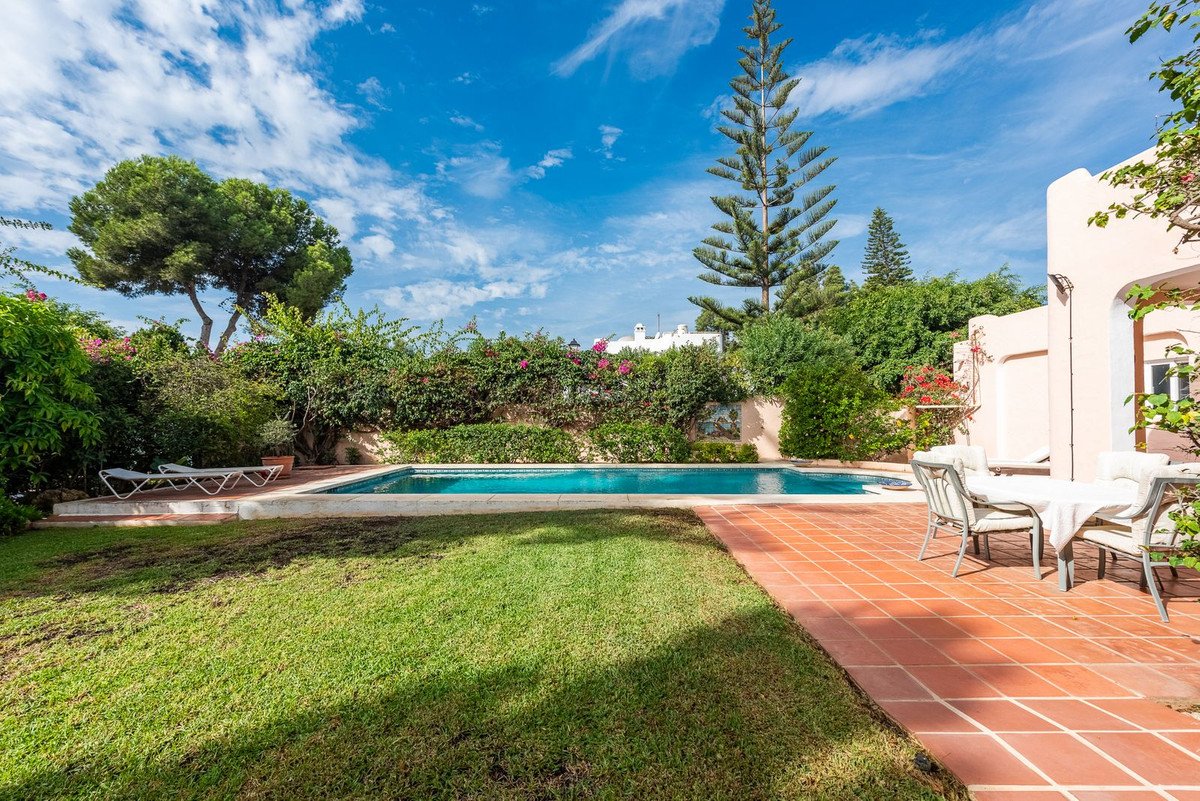 A swimming pool and patio with lounge chairs and an outdoor dining set, surrounded by lush greenery under a blue sky.