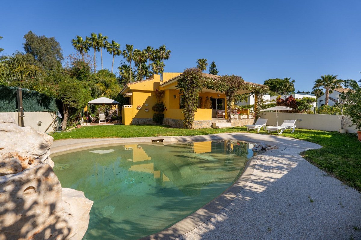 Yellow villa with a swimming pool, sun loungers, and lush garden with palm trees under a clear blue sky.