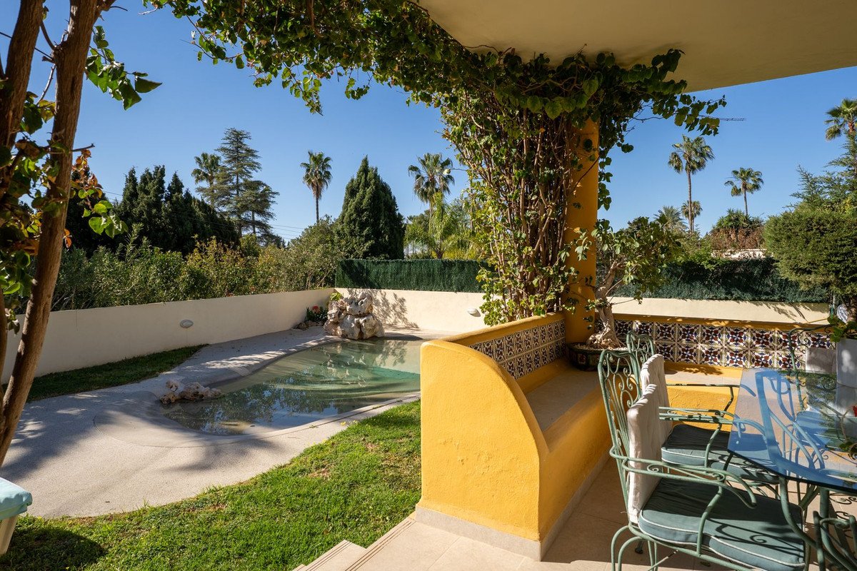 A covered patio with a built-in bench overlooks a pool and garden with palm trees and lush greenery under a clear blue sky.