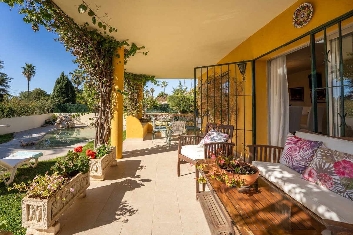 A covered patio with seating, overlooking a swimming pool and lush garden with palm trees under a clear blue sky.