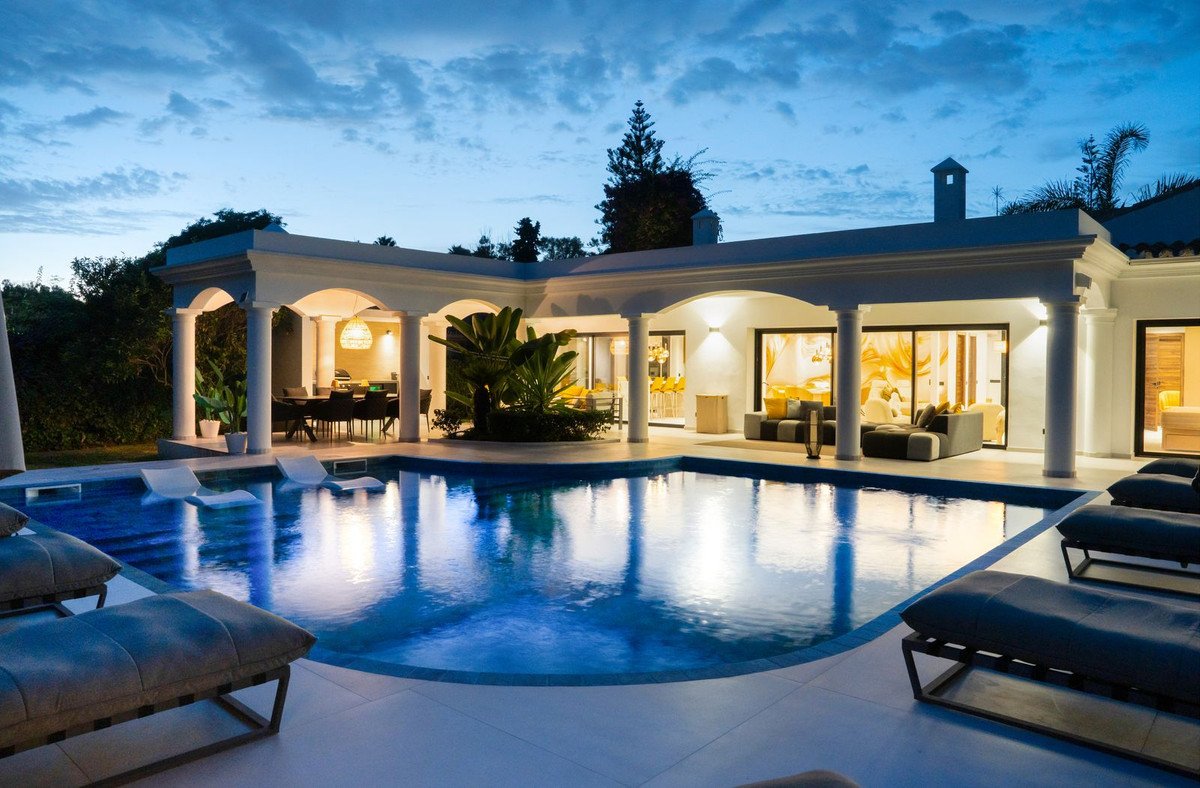 Luxurious villa with illuminated swimming pool and outdoor lounge area at dusk, featuring covered terrace and dining space.