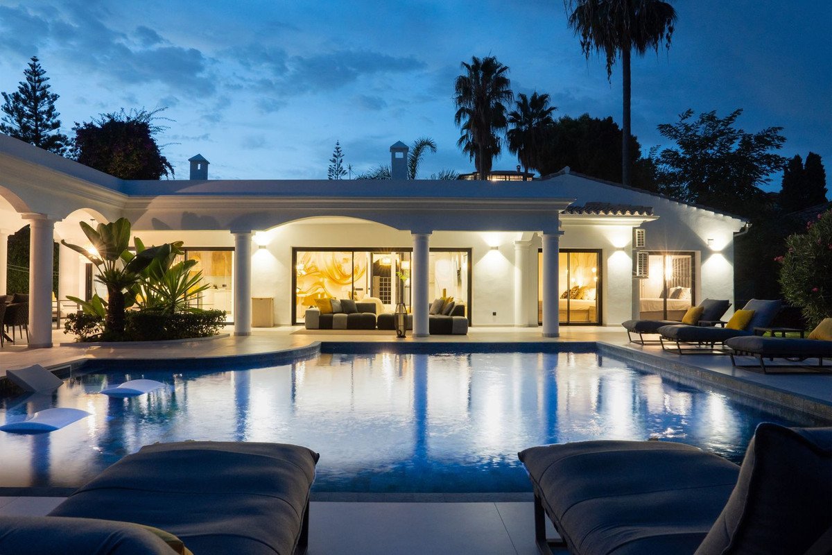 Luxurious villa at dusk with illuminated swimming pool, outdoor lounge seating, and tropical palm trees.