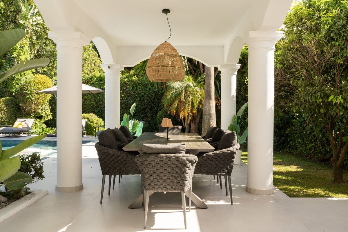 Covered outdoor dining area with a large table and chairs, overlooking a swimming pool and lush green gardens.