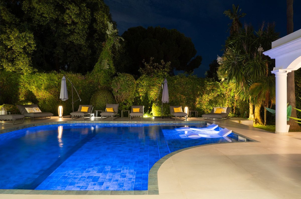 Luxurious outdoor swimming pool with illuminated water and lounge chairs by lush greenery at night.