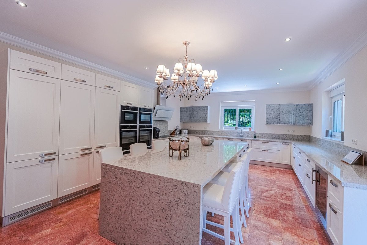 Spacious modern white kitchen with a large island, white bar stools, a chandelier, and integrated appliances. View of the garden through the window.