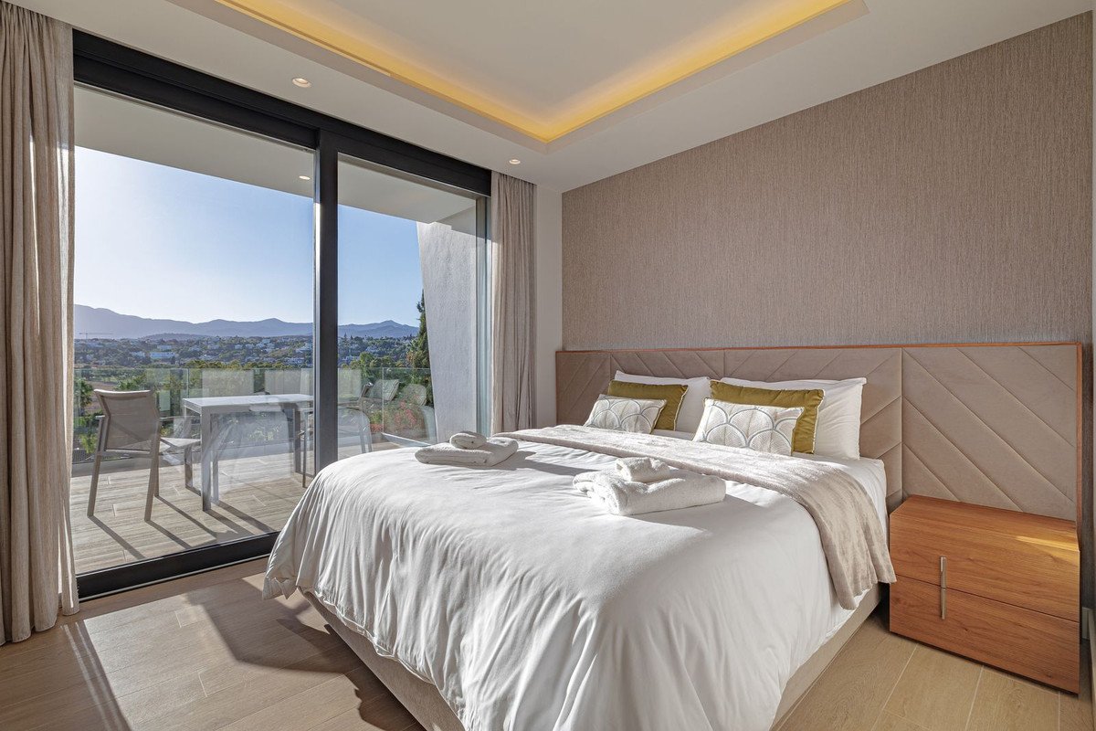 A bedroom with a large bed, overlooking a balcony with a sea view and distant hills.