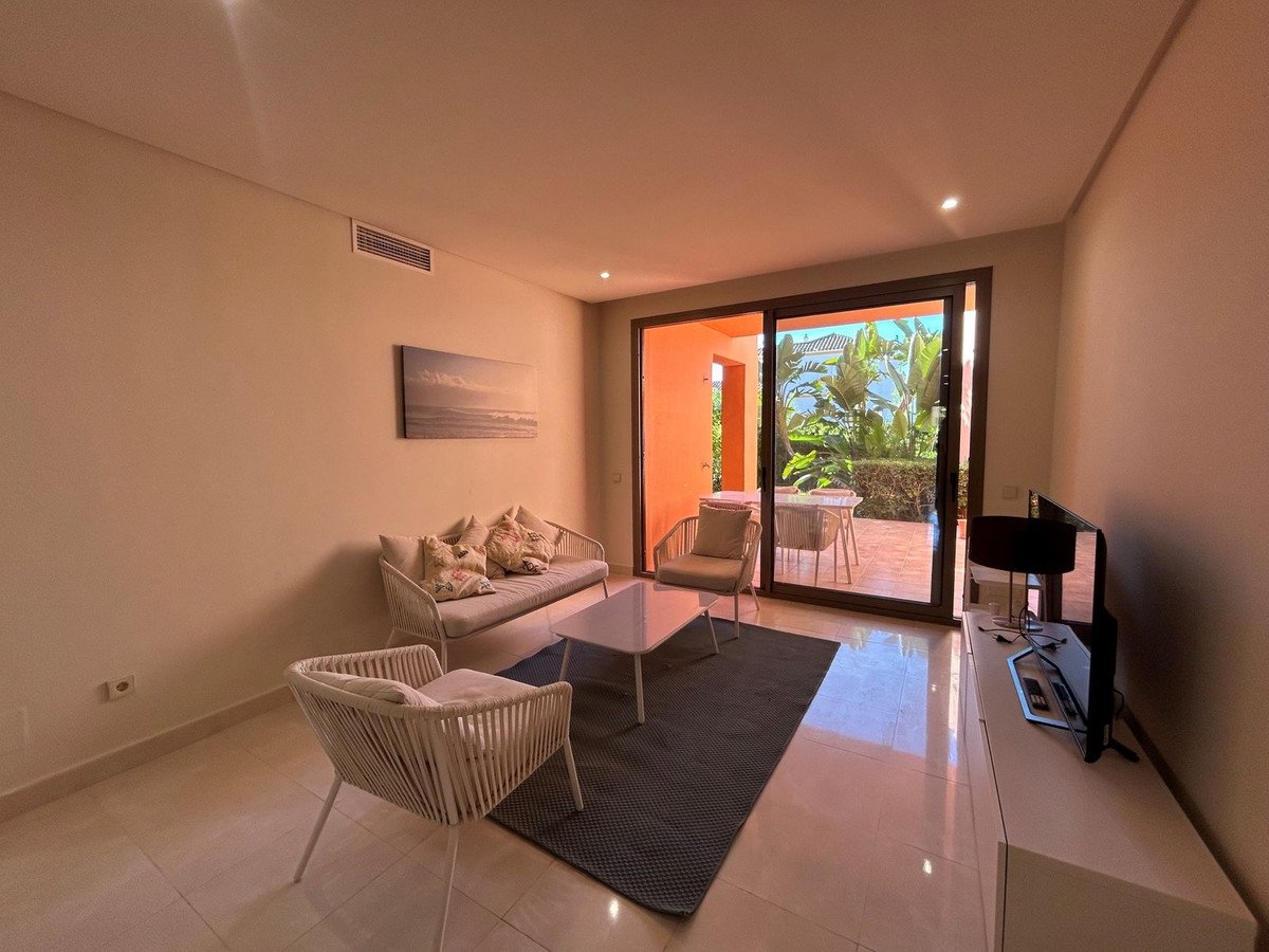 Modern living room with sofa, chairs, coffee table, and a TV, opening to a sunny garden terrace with outdoor seating.