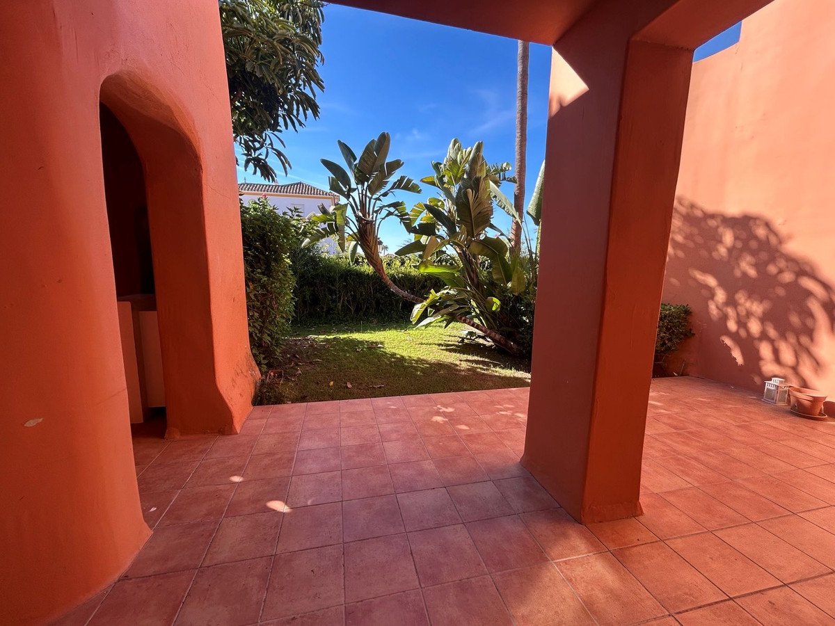 A tiled terrace with an archway leading to a green garden with palm trees under a blue sky.
