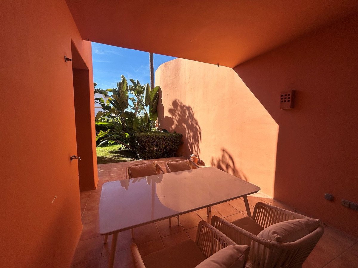 A covered terrace with a dining table and chairs, overlooking a lush garden and a bright blue sky.